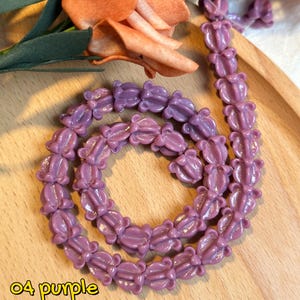 May include: A strand of purple flower-shaped beads. The beads are arranged in a circular pattern on a wooden tray. The text "04 purple" is in the lower left corner. The text below reads "Not all crown flowers beads are solid-colored - some come with beautiful gradient shades oh~!"