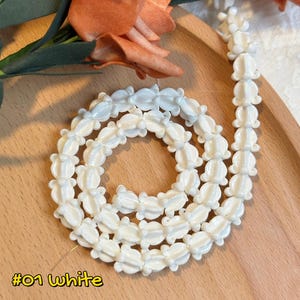 May include: White flower-shaped beads strung together, forming a necklace or strand. The beads are arranged in a spiral on a wooden surface. The image includes text that reads "#01 White" and a description about the beads.