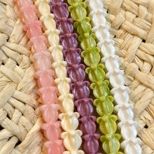 May include: Close-up of several strands of colorful beads. The beads are shaped like flowers and come in shades of pink, cream, purple, green, and white. The background is a woven straw material.
