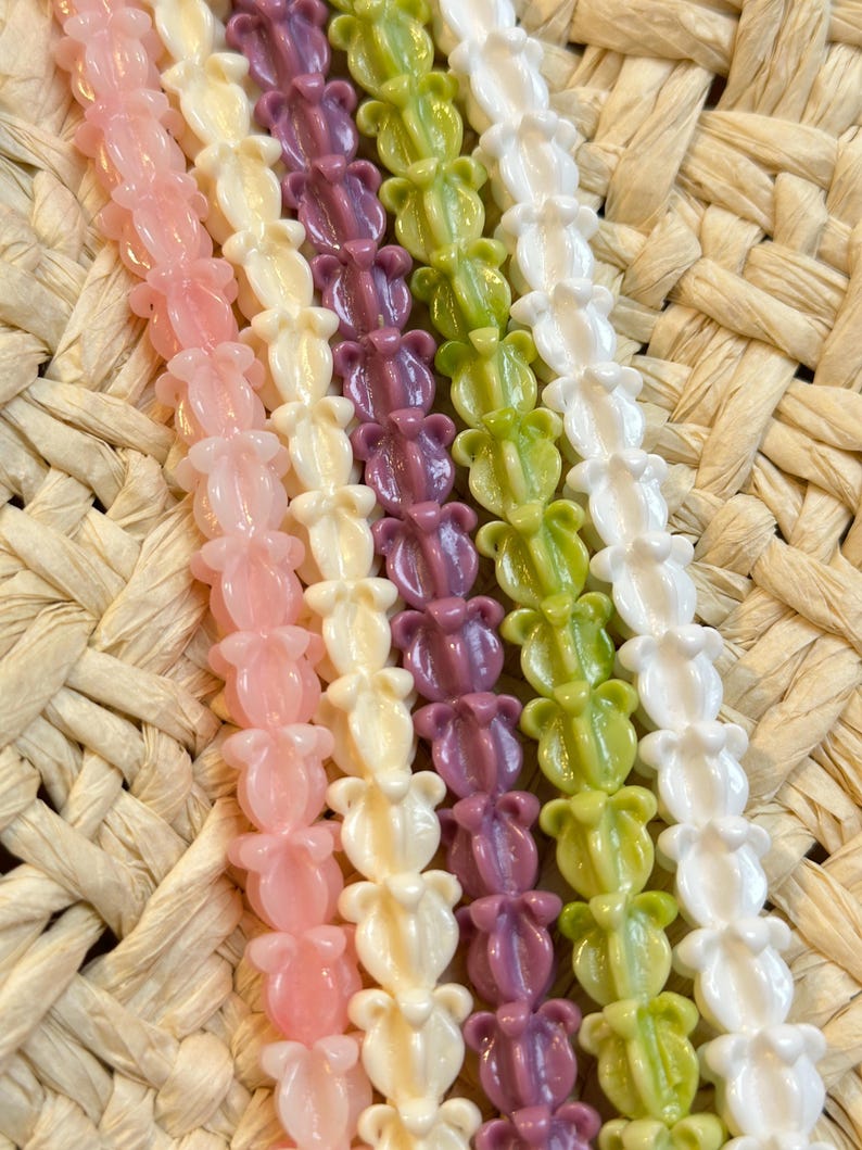 May include: Close-up of several strands of colorful beads. The beads are shaped like flowers and come in shades of pink, cream, purple, green, and white. The background is a woven straw material.