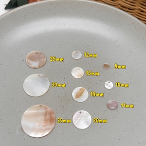 May include: A collection of round, flat shell discs with small holes for hanging, labeled with sizes ranging from 8mm to 35mm. The discs feature iridescent, pearlescent surfaces in shades of white, cream, and light brown, arranged in a grid.