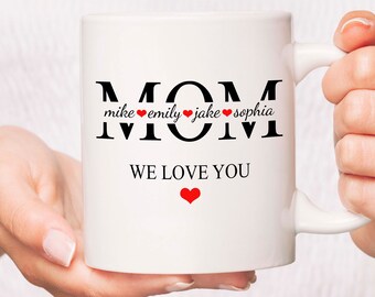 Könnte beinhalten: Weiße Keramiktasse mit schwarzem Text, der "MOM" lautet, mit den Namen "mike", "emily", "jake" und "sophia", die unter dem "M" mit einem roten Herzen zwischen jedem Namen geschrieben sind. Der Text "WE LOVE YOU" steht unter den Namen mit einem roten Herzen unter dem Text.