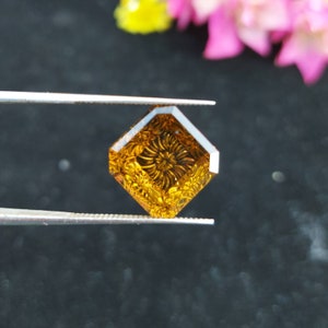 May include: A square-shaped, carved gemstone with a floral design. The gemstone is a deep amber color and is being held by tweezers.