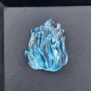 May include: A blue aquamarine gemstone carved into the shape of a flame. The gemstone is translucent and has a smooth, polished finish.