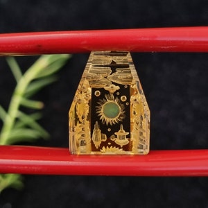 May include: A clear, rectangular, yellow-tinted crystal with etched designs of clouds, sun, and buildings. The crystal is held by red chopsticks, with green foliage in the background. The crystal has a central sun design with a green center.