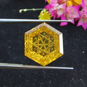 May include: A yellow hexagonal gemstone with a detailed star-shaped design in the center. The gemstone is held by a pair of tweezers.