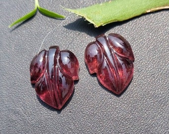 May include: Two polished, leaf-shaped gemstones in a deep burgundy hue. The gems have carved details mimicking leaf veins. A green leaf and a sprig of grass are in the background, set against a dark, textured surface.