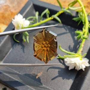 May include: A brown, faceted gemstone pendant with a carved floral design. The pendant is held by tweezers against a black background.