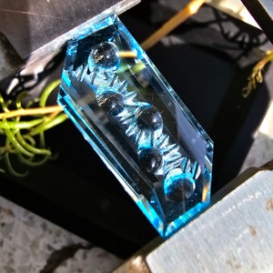 May include: A rectangular pendant made of clear blue glass with a row of black beads inside. The beads are set in a decorative pattern of small points.