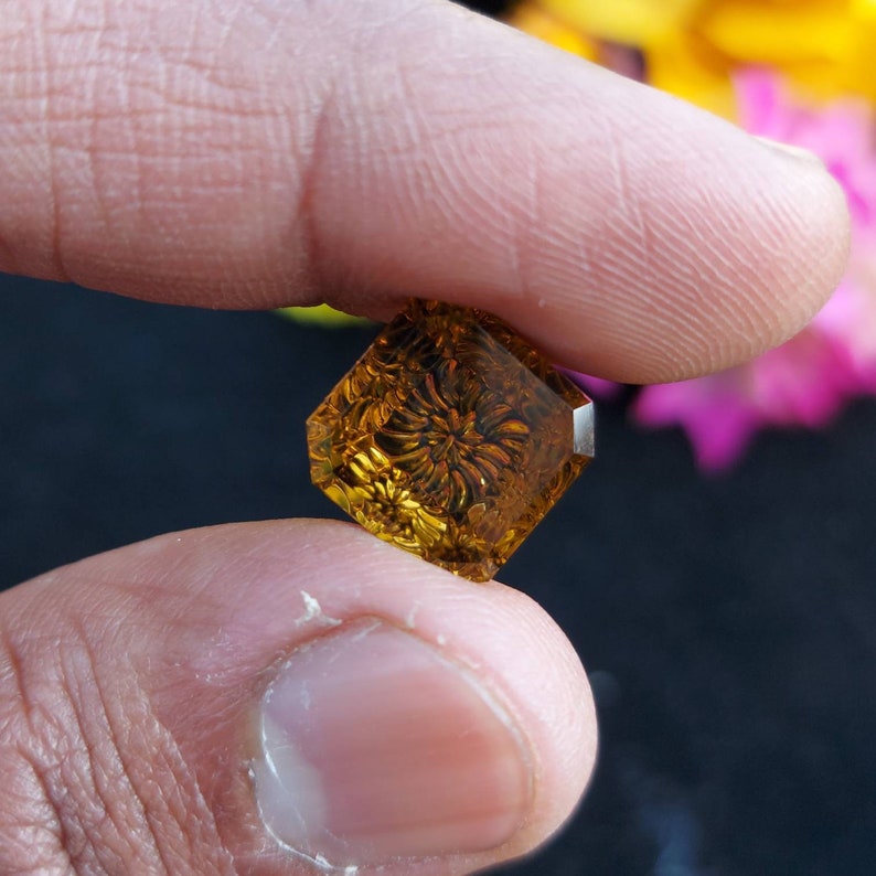 May include: A square-shaped, carved gemstone with a floral design. The gemstone is a deep amber color and is translucent.