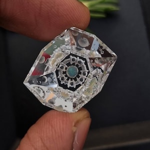 Könnte beinhalten: Ein klarer Kristall mit geometrischer Form und einem grünen Edelstein in der Mitte. Der Kristall hat ein detailliertes Design mit kleinen weißen Punkten und einem kreisförmigen Muster.
