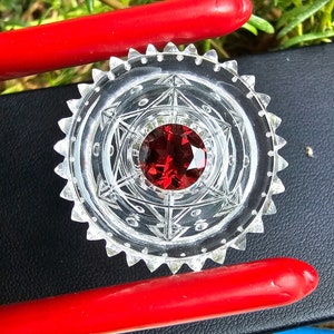 May include: A clear glass circular pendant with a red gemstone in the center. The pendant has a geometric design with a starburst pattern.