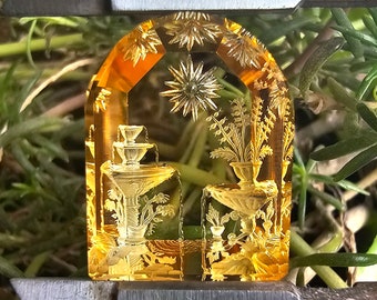 Può includere: Un cammeo in vetro giallo con un design intricato di una fontana e fiori. Il cammeo è incastonato in una cornice di metallo ed è circondato da fogliame verde.