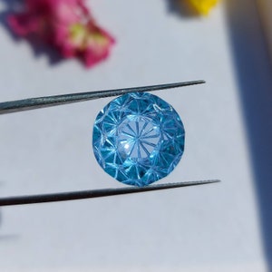May include: A blue gemstone with a faceted cut, held by tweezers. The gemstone is round and has a starburst pattern in the center.