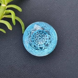 Könnte beinhalten: Ein runder, hellblauer Glasgegenstand mit einem detaillierten floralen Muster, das in seine Oberfläche geätzt ist. Das Objekt befindet sich vor einem dunklen Hintergrund, mit einem Zweig grünen Laubes in der oberen linken Ecke. Das Glas hat ein facettiertes Aussehen.