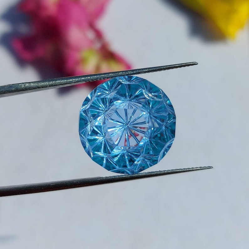 May include: A blue gemstone with a faceted cut, held in tweezers. The gemstone has a star-like pattern in the center.