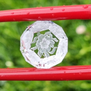 May include: A clear glass paperweight with a carved floral design. The paperweight is octagonal in shape and has a raised, detailed flower design in the center.