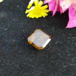 May include: A square, faceted, amber-colored gemstone with a floral pattern inside. The gemstone is set against a black background.
