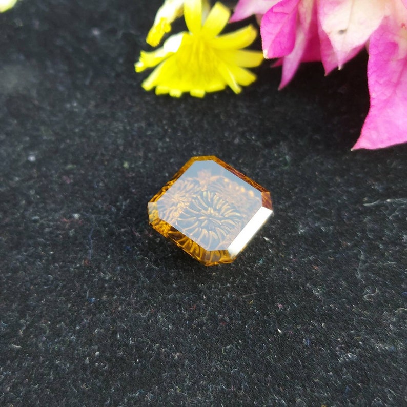 May include: A square, faceted, amber-colored gemstone with a floral pattern inside. The gemstone is set against a black background.