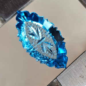 May include: A blue faceted oval-shaped gemstone with a carved design. The gemstone is set in a metal frame.