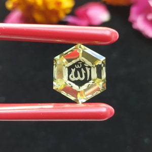 May include: A hexagonal, yellow-toned crystal pendant with the Arabic inscription "Allah" etched in the center. The pendant is held by red tweezers against a dark background, with blurred flowers in the background.