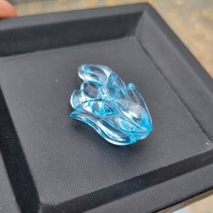 May include: A carved blue topaz gemstone in the shape of a flower. The gemstone is on a black surface.