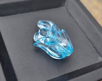 Puede incluir: Una piedra preciosa de topacio azul tallada en forma de flor. La piedra preciosa está sobre una superficie negra.