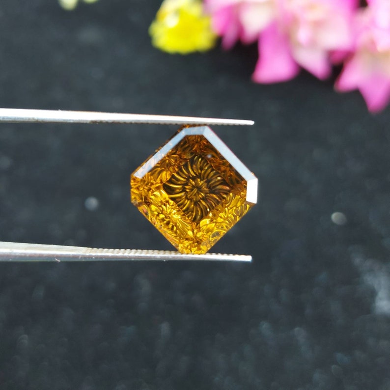 May include: A square-shaped, carved gemstone with a floral design. The gemstone is a deep amber color and is being held by tweezers.