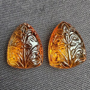 May include: Two carved amber-colored glass pendants with a floral design. The pendants are shaped like a teardrop and have a detailed floral pattern.