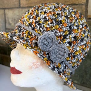 Puede incluir: Un sombrero cloche de ganchillo en una mezcla de colores que incluye amarillo, naranja, negro, blanco y gris. El sombrero presenta un ala pequeña y un detalle de lazo gris en el lateral. El sombrero se muestra en una cabeza de maniquí.