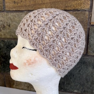 Op de afbeelding: Een lichtbruine gehaakte beanie-muts met een open weefpatroon. De hoed wordt getoond op een mannequinhoofd met rode lippenstift en geschilderde wangen. De hoed heeft een neutrale kleur en is geschikt voor elk seizoen.