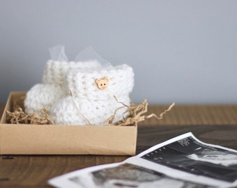 Peut inclure: Petites chaussons de bébé en crochet blanc avec un petit bouton en forme d'ours, dans une boîte brune avec du papier déchiqueté, à côté d'une image d'échographie en noir et blanc.