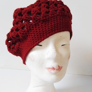 May include: A red crocheted beret with a lacy design. The beret is worn on a white mannequin head.