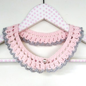 May include: A pink and gray crocheted collar with a pearl button in the center. The collar is displayed on a white polka dot hanger.