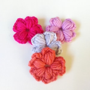 May include: Four crocheted flowers in different colors: pink, gray, coral, and a brighter pink. The flowers are all the same size and shape, with five petals each.