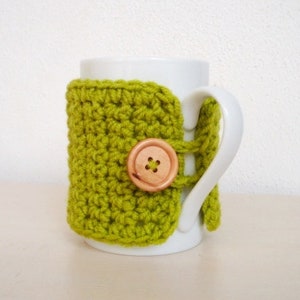 May include: A white ceramic mug with a green crocheted cozy. The cozy has a wooden button.