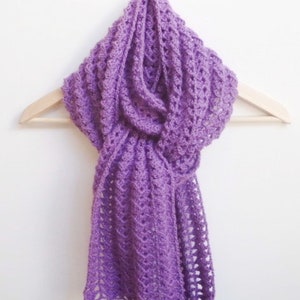 Puede incluir: Una bufanda de ganchillo de color morado con un delicado patrón de calado. La bufanda está colgada sobre una percha de madera.