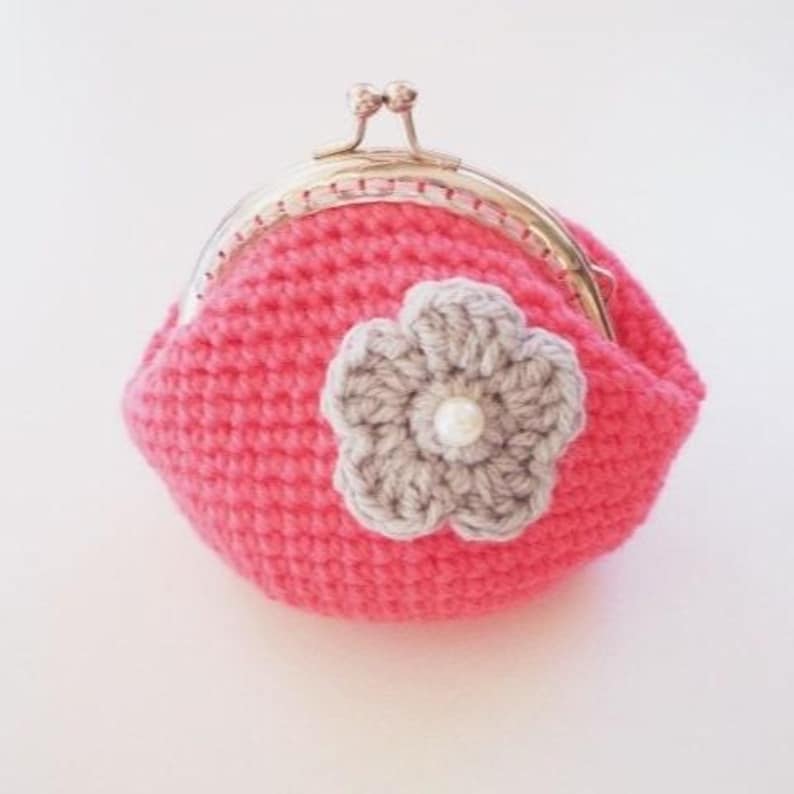 May include: A bright pink crocheted coin purse featuring a metallic gold-tone kiss-lock clasp. The front of the purse is decorated with a light gray crocheted flower applique, which has a small white pearl bead centered in the middle of the petals.