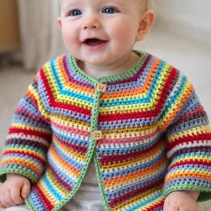 May include: A smiling baby wearing a colorful, striped crochet cardigan with wooden buttons. The cardigan features a rainbow of colors, including red, orange, yellow, green, blue, and purple. The baby is wearing gray pants.