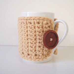 May include: A white ceramic mug with a beige crocheted cozy and a brown button. The cozy is crocheted in a simple stitch pattern.