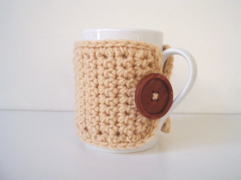 May include: A white ceramic mug with a beige crocheted cozy and a brown button. The cozy is crocheted in a simple stitch pattern.