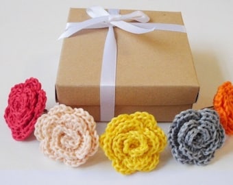 May include: Five crocheted flower rings in a variety of colors, including red, pink, yellow, gray, and orange, are arranged in a row on a white surface. The rings are packaged in a brown gift box with a white ribbon.