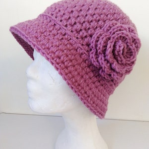 May include: A handmade, lavender-colored crochet hat with a small brim and a decorative flower on the side. The hat is made of a textured yarn, and the flower is made of the same material.
