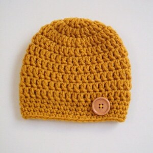 May include: A mustard yellow beanie crafted with a thick crochet stitch pattern. The hat features a rounded crown and a single, circular, light brown four-hole button securely attached to the lower right side of the front panel.