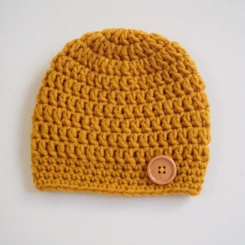May include: A mustard yellow beanie crafted with a thick crochet stitch pattern. The hat features a rounded crown and a single, circular, light brown four-hole button securely attached to the lower right side of the front panel.