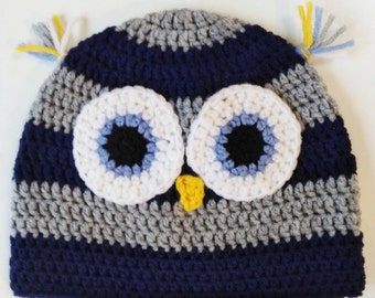 May include: A blue, gray, and white striped crocheted owl hat with yellow yarn accents. The hat has large white eyes with blue and black details.