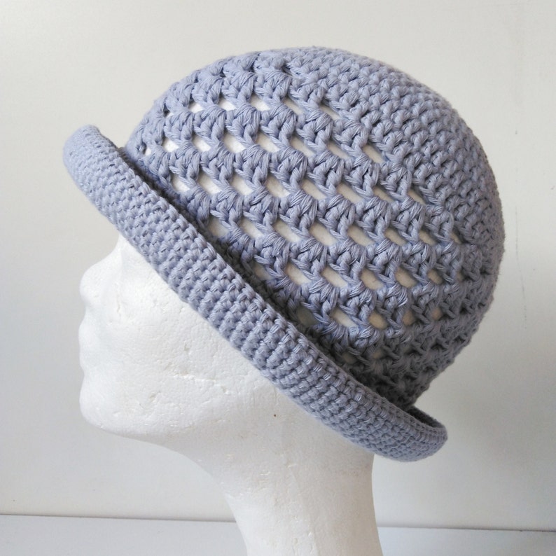 Peut inclure: Un chapeau crochet&eacute; gris clair avec un large bord et un motif en tissage ouvert. Le chapeau est sur une t&ecirc;te de mannequin blanche.