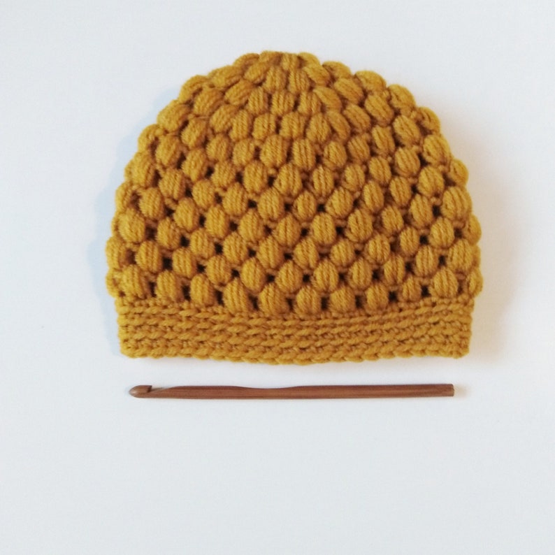 May include: A mustard yellow crocheted beanie hat with a textured bobble stitch pattern. The hat is made with a single crochet stitch and has a simple brim.