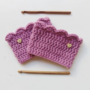 Könnte beinhalten: Zwei lavendelfarbene Häkel-Handgelenkmanschetten mit gewelltem Rand und herzförmigen Holzknöpfen. Zwei Häkelnadeln aus Holz sind über und unter den Manschetten auf einer weißen Oberfläche positioniert. Die Manschetten sind handgefertigt.
