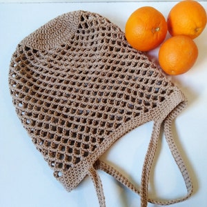 May include: A tan crocheted market bag with a net-like design, featuring two handles. Three oranges are placed next to the bag on a white surface. The bag is designed for carrying groceries or other items.